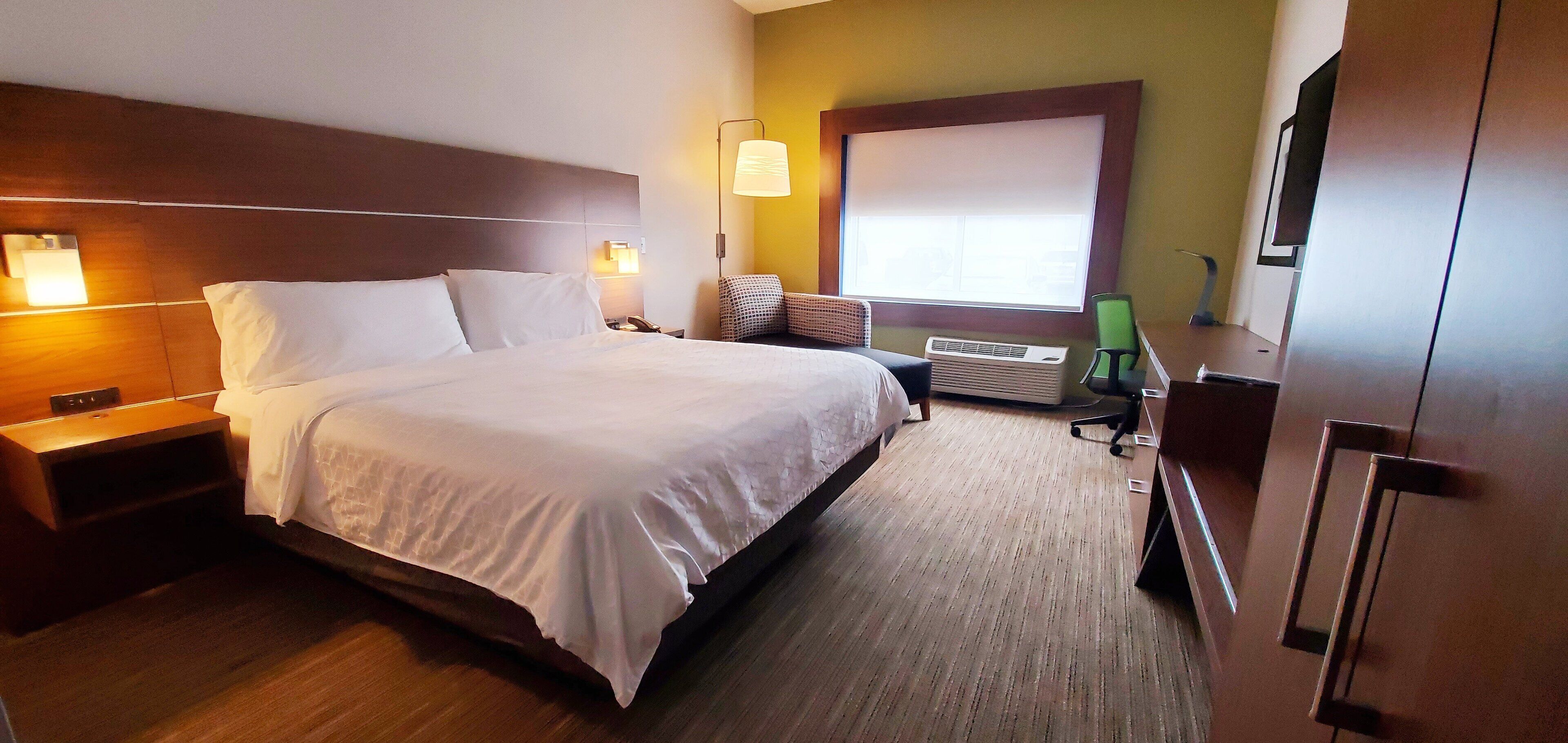 Habitación de hotel con una cama grande, lámparas de noche, un escritorio, una silla, un equipo de aire acondicionado y una ventana con persianas.