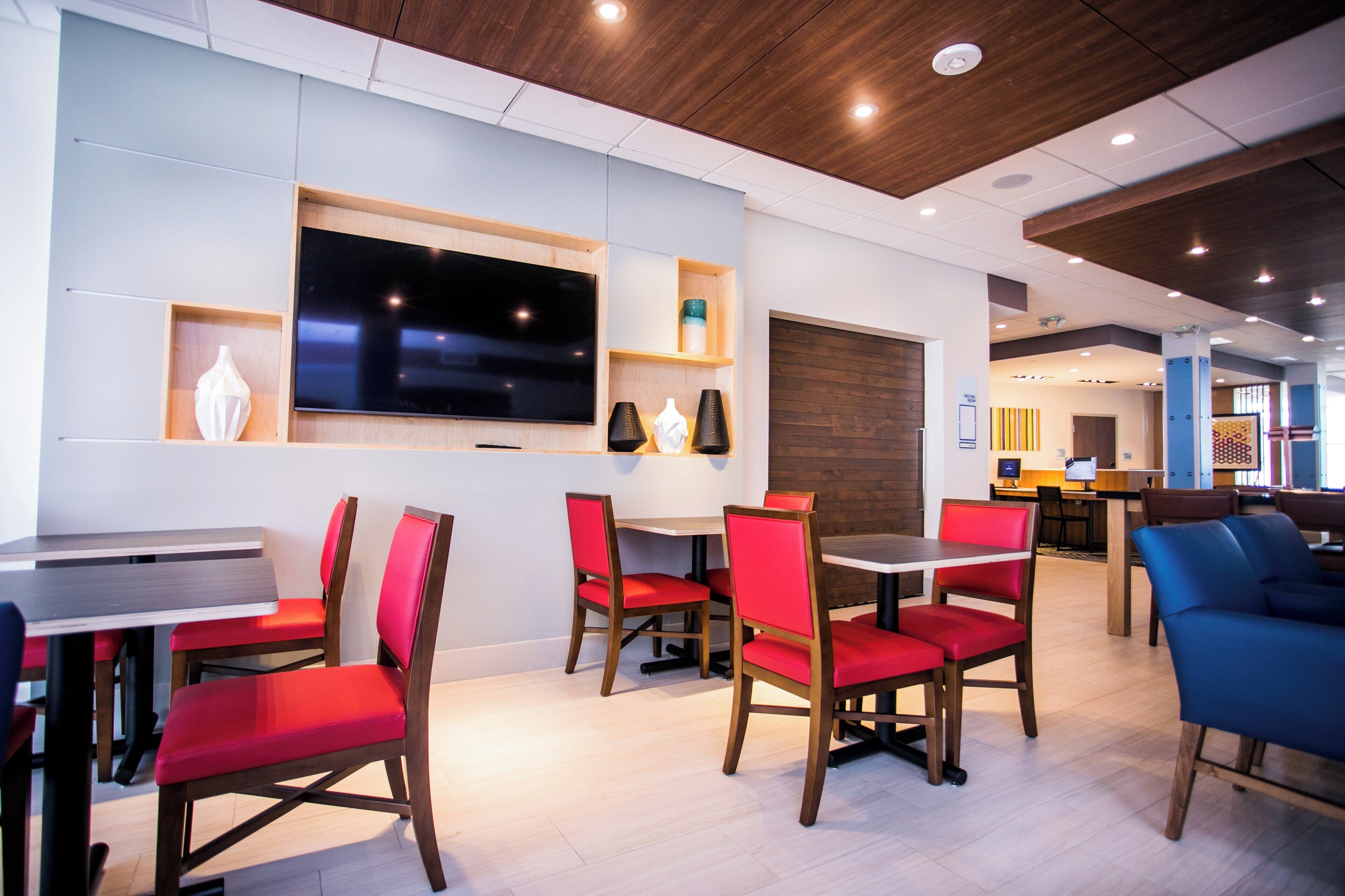 A modern hotel lobby with a flat-screen TV, wooden furniture, and a mix of red and blue chairs.