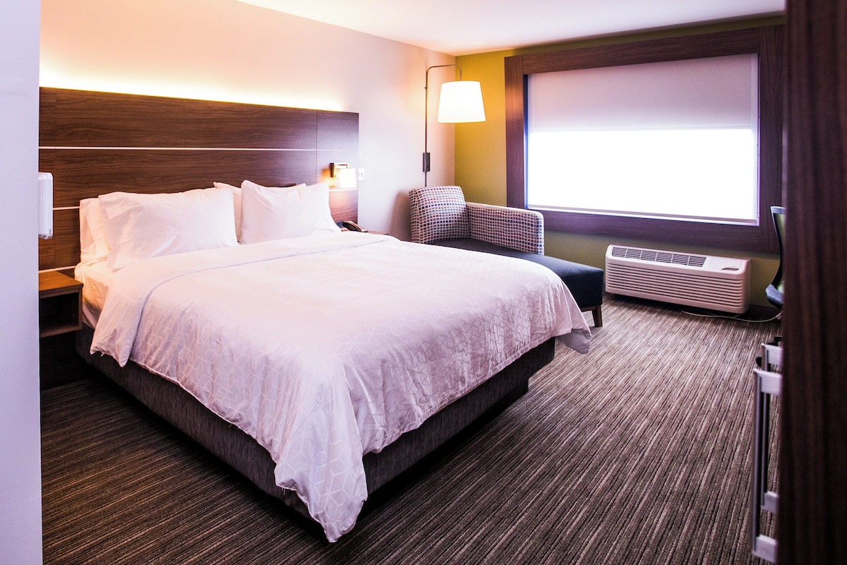 A hotel room with a large bed, a chair, a desk, a lamp, and a window.