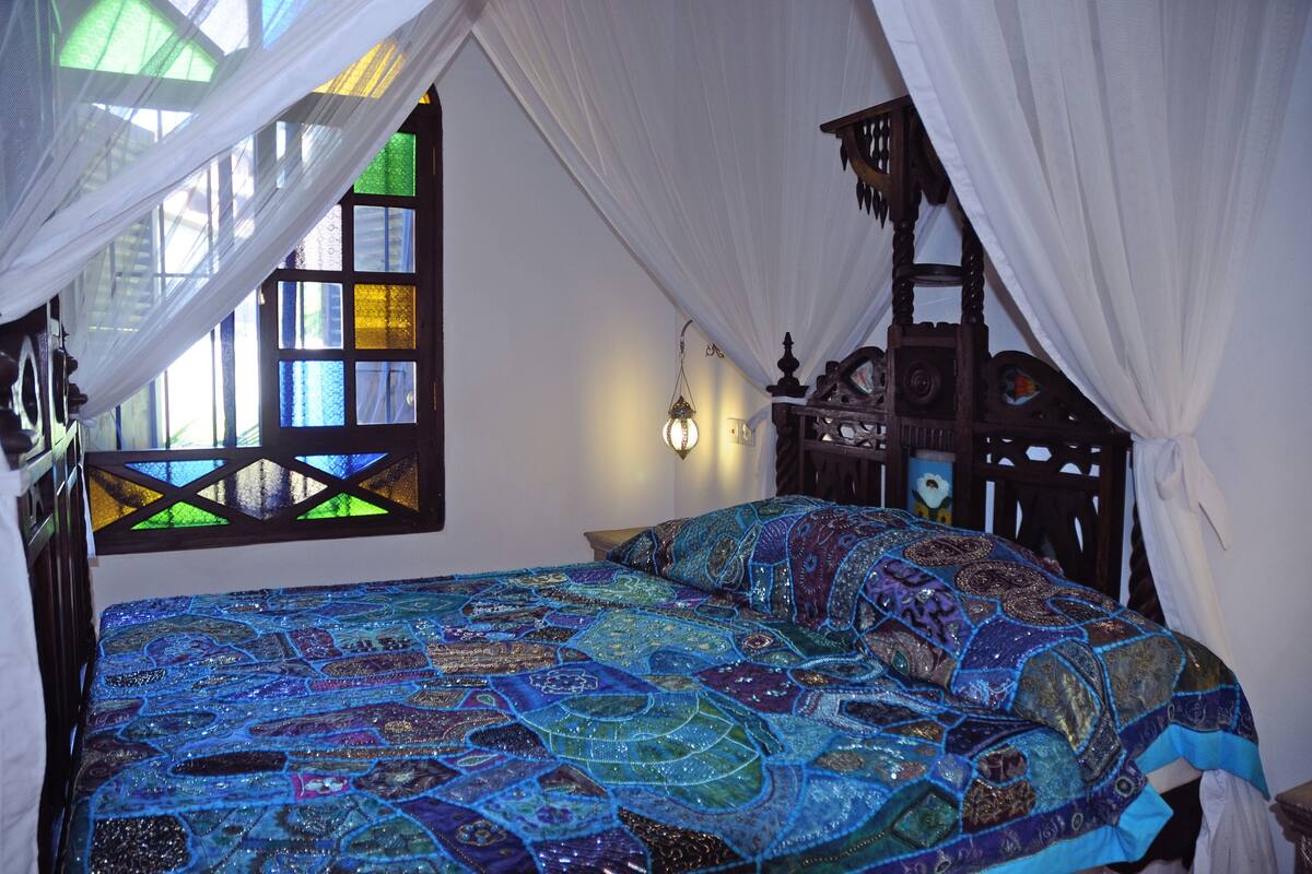 A bed with a colorful quilt, a window with stained glass, and a wooden headboard.