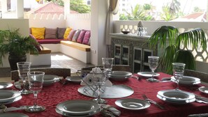 Uma sala de jantar com toalha vermelha, preparada para uma refeição, com pratos, copos e talheres. Há um sofá com almofadas, um vaso de planta e vista para palmeiras e edifícios lá fora.