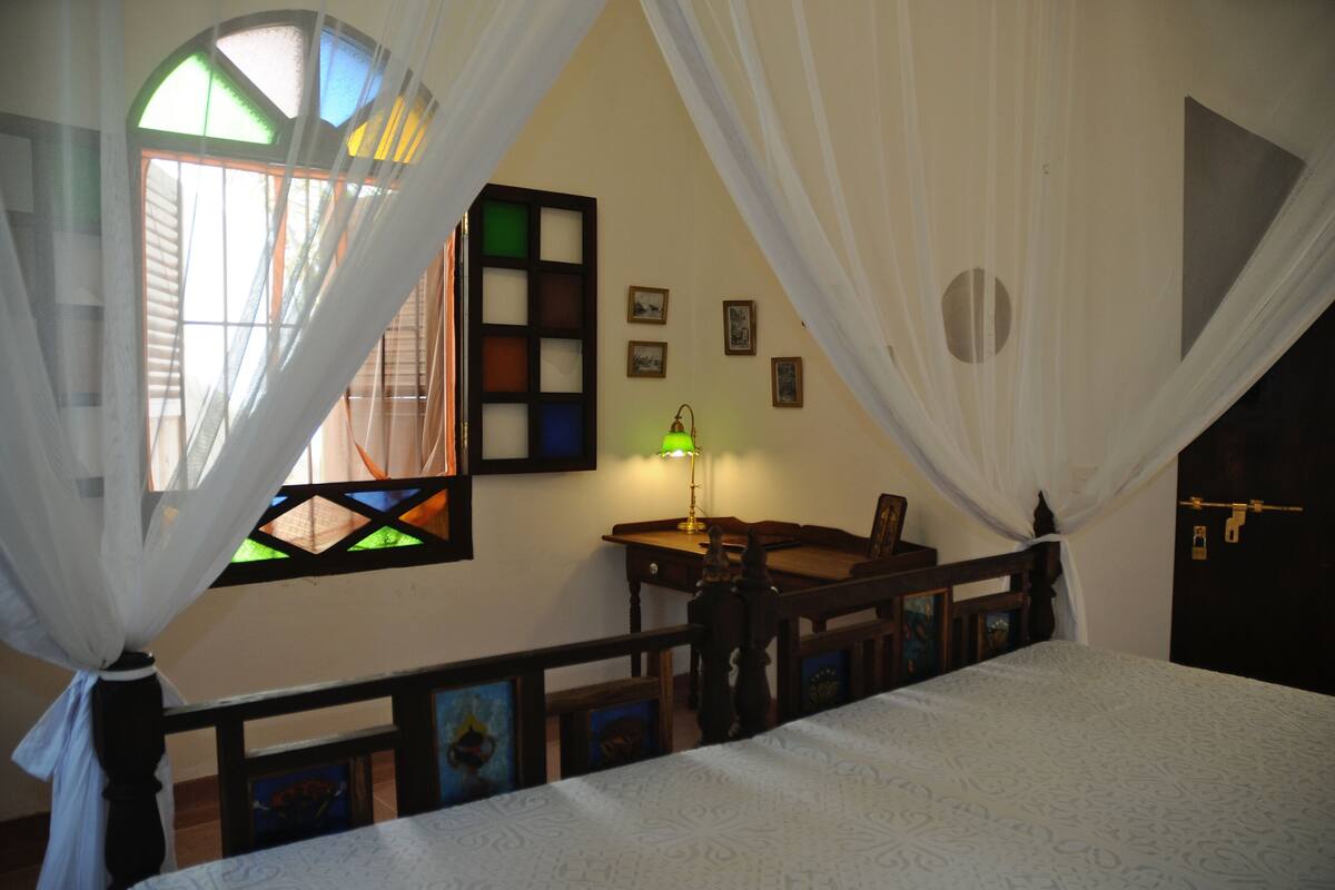A bedroom with a bed, a nightstand, a lamp, and a window with a stained glass design.