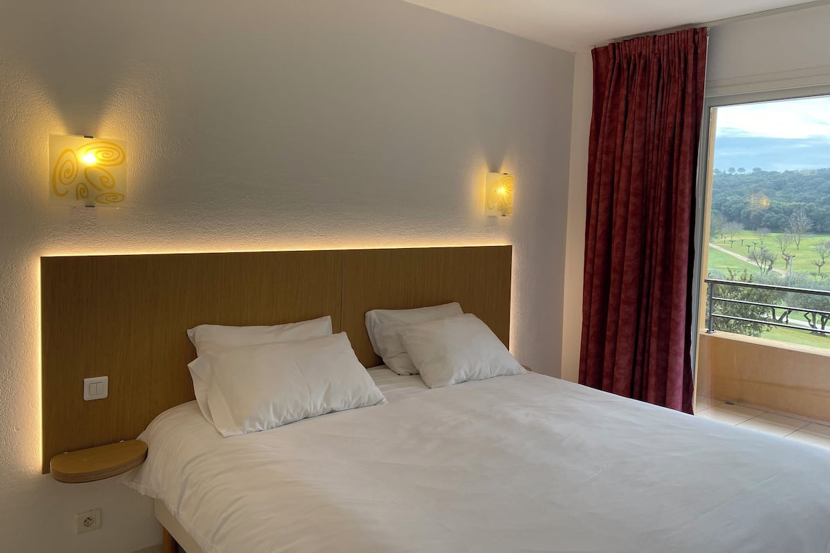 A hotel room with a large bed, two wall-mounted lights, a wooden headboard, a window with a view of a landscape, and a red curtain.