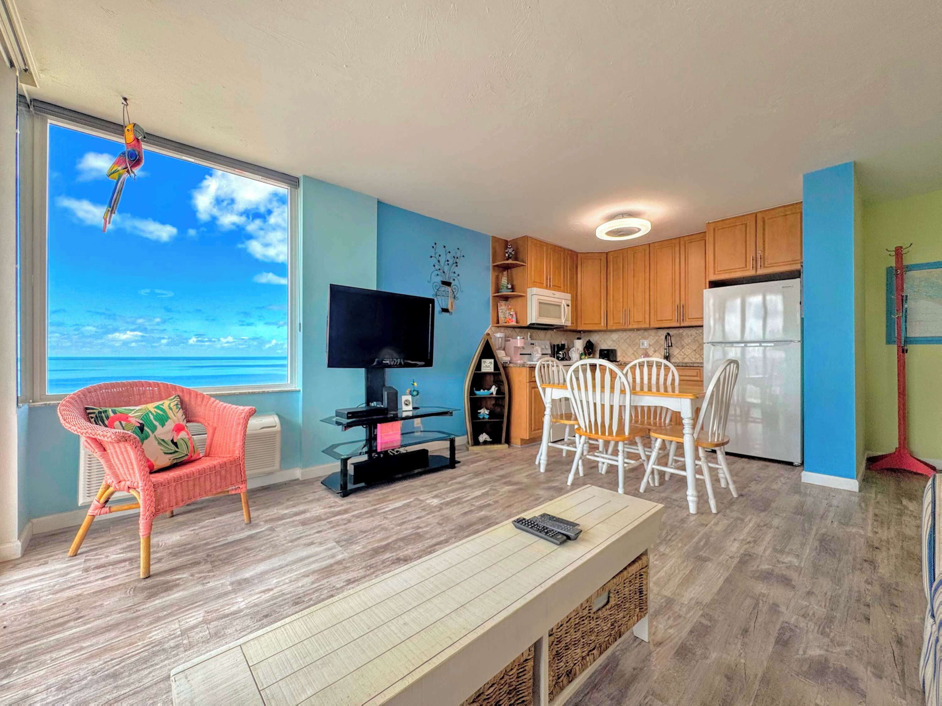 A living room with a TV, a dining area with chairs, a kitchen with cabinets, and a view of the ocean.