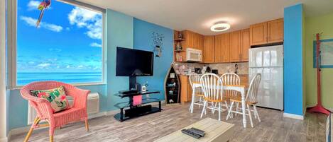 A living room with a TV, a dining area with chairs, a kitchen with cabinets, and a view of the ocean.