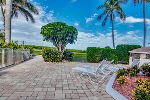 A paved patio with lounge chairs, a large tree, and a view of a golf course.