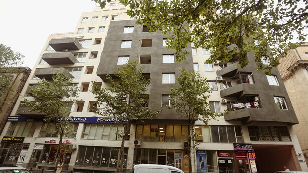 A multi-story building with balconies, storefronts, and parked cars. - One Bedroom apartment for shopaholics (Tbilisi)