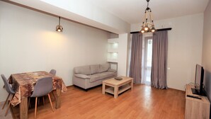 A living room with a sofa, a dining table with chairs, a coffee table, and a TV stand. - One Bedroom apartment for shopaholics (Tbilisi)
