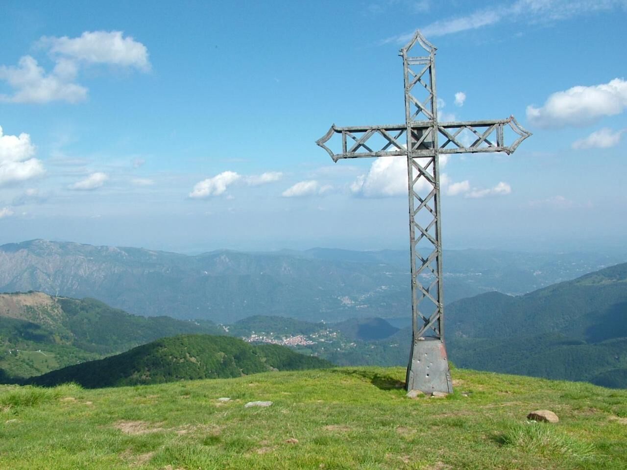 Una estructura metálica en forma de cruz sobre una colina cubierta de hierba, con una vista panorámica de montañas y valles al fondo.
