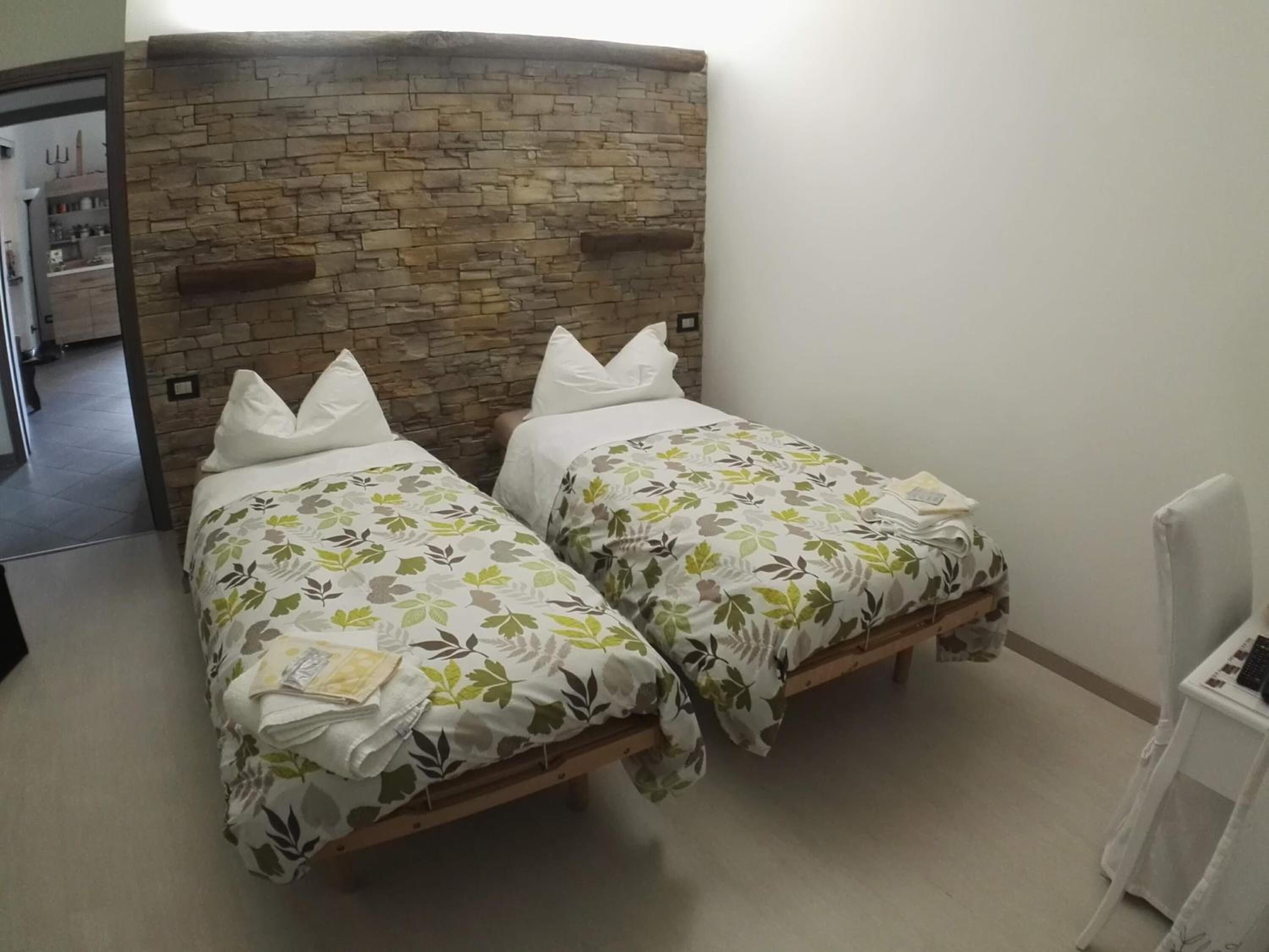 A room with two single beds, each with a floral-patterned bedspread and white pillows. The beds have wooden frames and are positioned against a wall with a stone headboard.