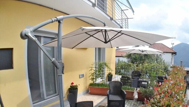 An outdoor patio area with wicker furniture, a large umbrella, and potted plants. - B&B Guglielmina (Quarna Sotto)