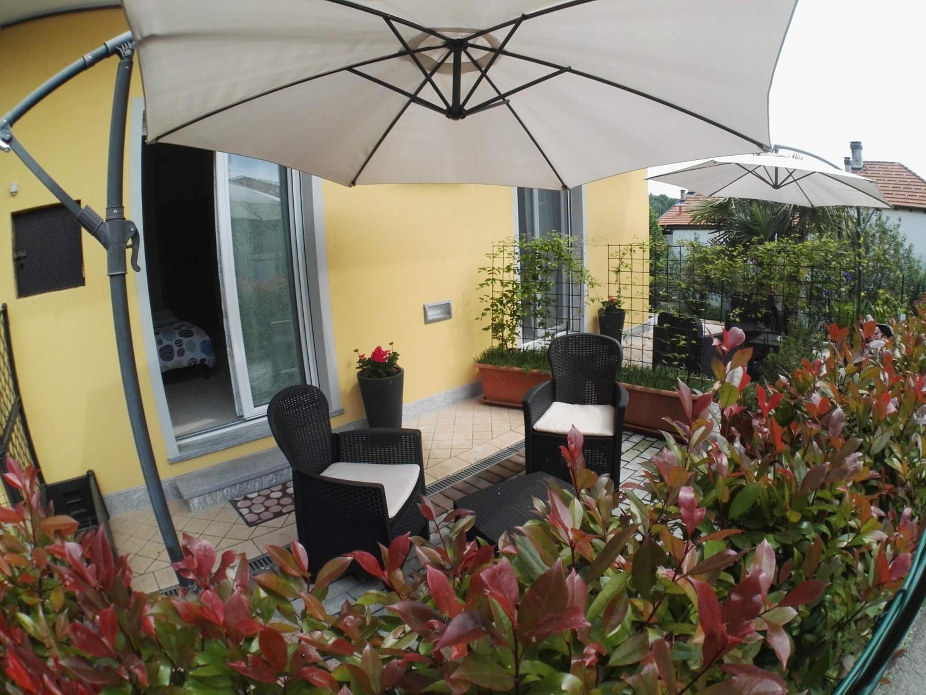 Ein möblierter Patio-Bereich im Freien mit zwei schwarzen Korbstühlen, einem Tisch, einem großen Sonnenschirm und Topfpflanzen.