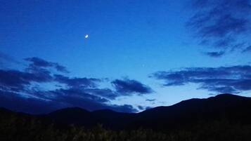 A night sky with a crescent moon and stars, over a mountainous landscape with sparse vegetation.