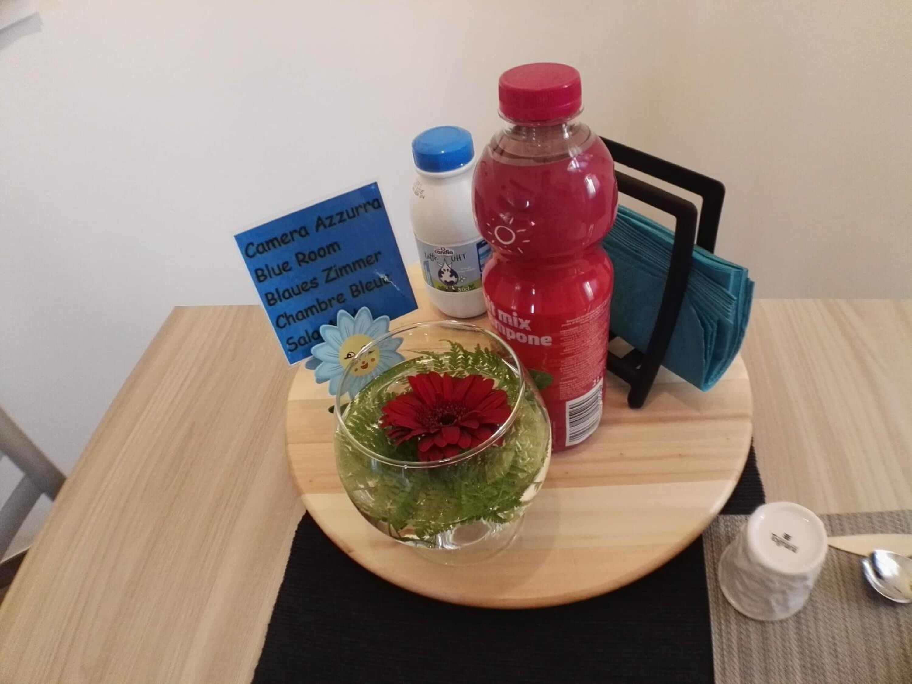 Une table avec un verre d’eau, une bouteille rouge, un petit récipient blanc et un panneau bleu avec du texte.