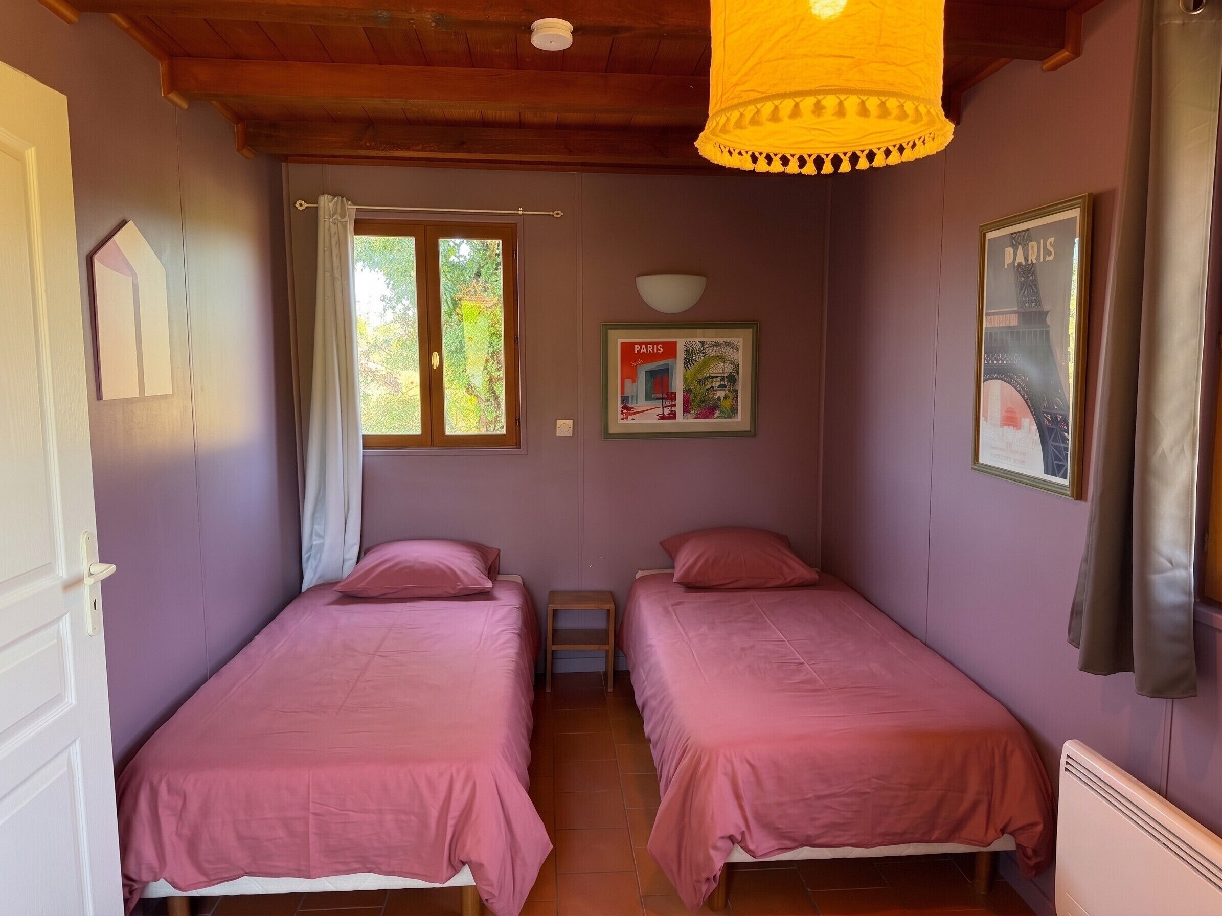 A room with two beds, a window with curtains, a radiator, and a poster of the Eiffel Tower.