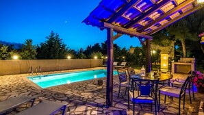 An outdoor pool area with stone flooring, a pergola with blue lighting, and a table with chairs. - Boschetto Holiday Village (Zakynthos)