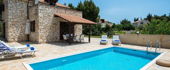 A stone building with a tiled roof, a swimming pool, lounge chairs, and a table with chairs.