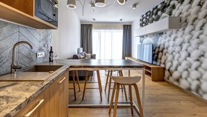 Une cuisine moderne dotée d’un plan de travail en marbre, de meubles en bois et d’un four encastré. On y trouve un coin repas avec une table et des chaises en bois, ainsi qu’une grande fenêtre agrémentée de rideaux.