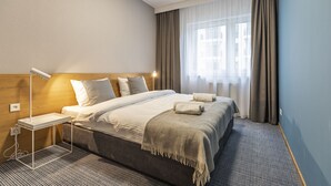 Une chambre d’hôtel bien aménagée, avec un grand lit, une table de chevet dotée d’une lampe et une vue sur la ville à travers de fines rideaux.