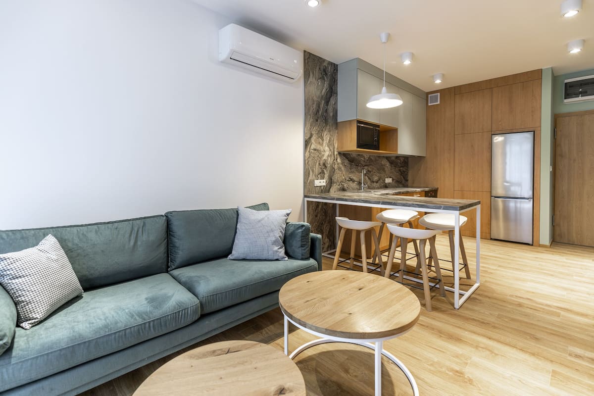 Una sala de estar moderna con un sofá verde, dos mesas redondas de madera y una zona de cocina con barra alta y taburetes.