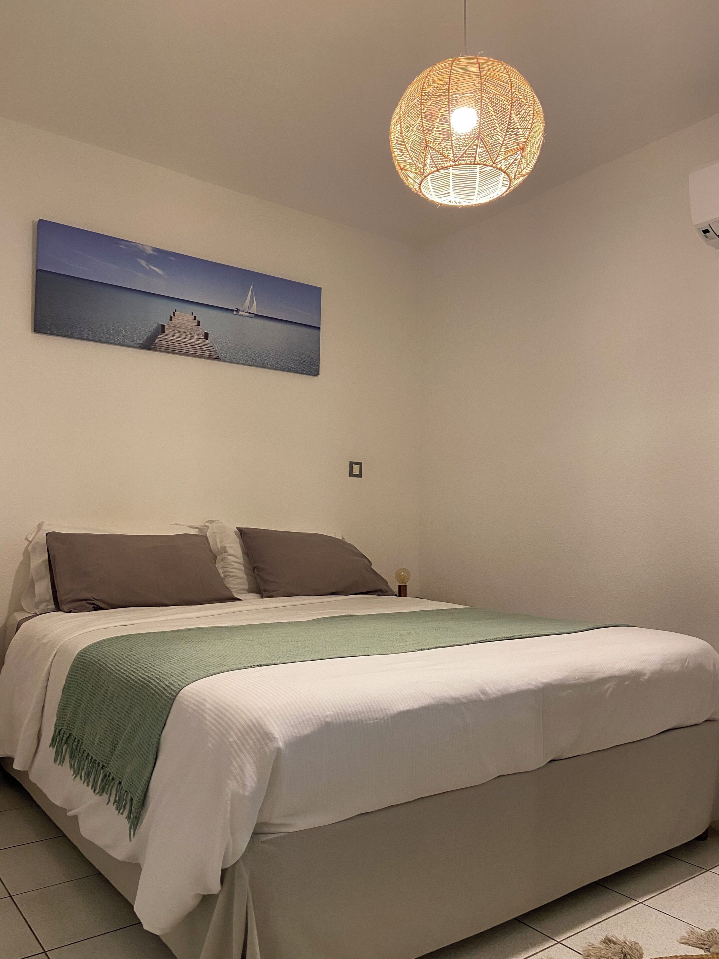 Une chambre à coucher avec un lit, des oreillers, un plaid vert, un tableau représentant un voilier accroché au mur et un luminaire suspendu.