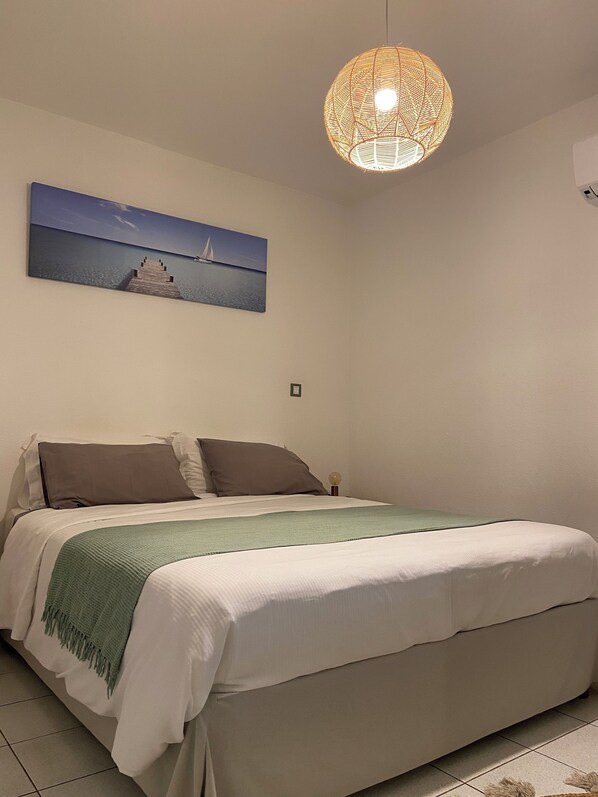 A bedroom with a bed, pillows, a green throw, a wall-mounted picture of a sailboat, and a hanging light fixture.