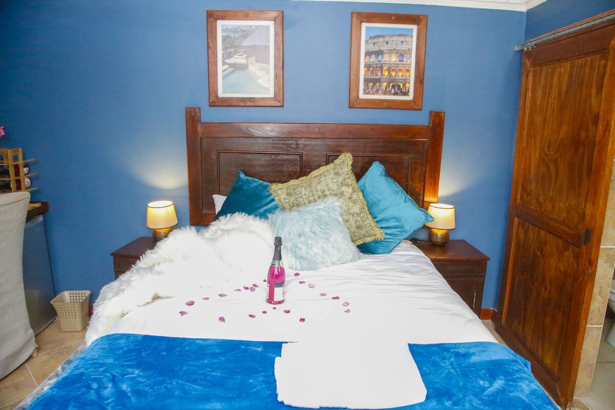 A neatly made bed with a blue blanket, white pillows, and a bottle of champagne. There are two framed pictures on the wall above the bed.