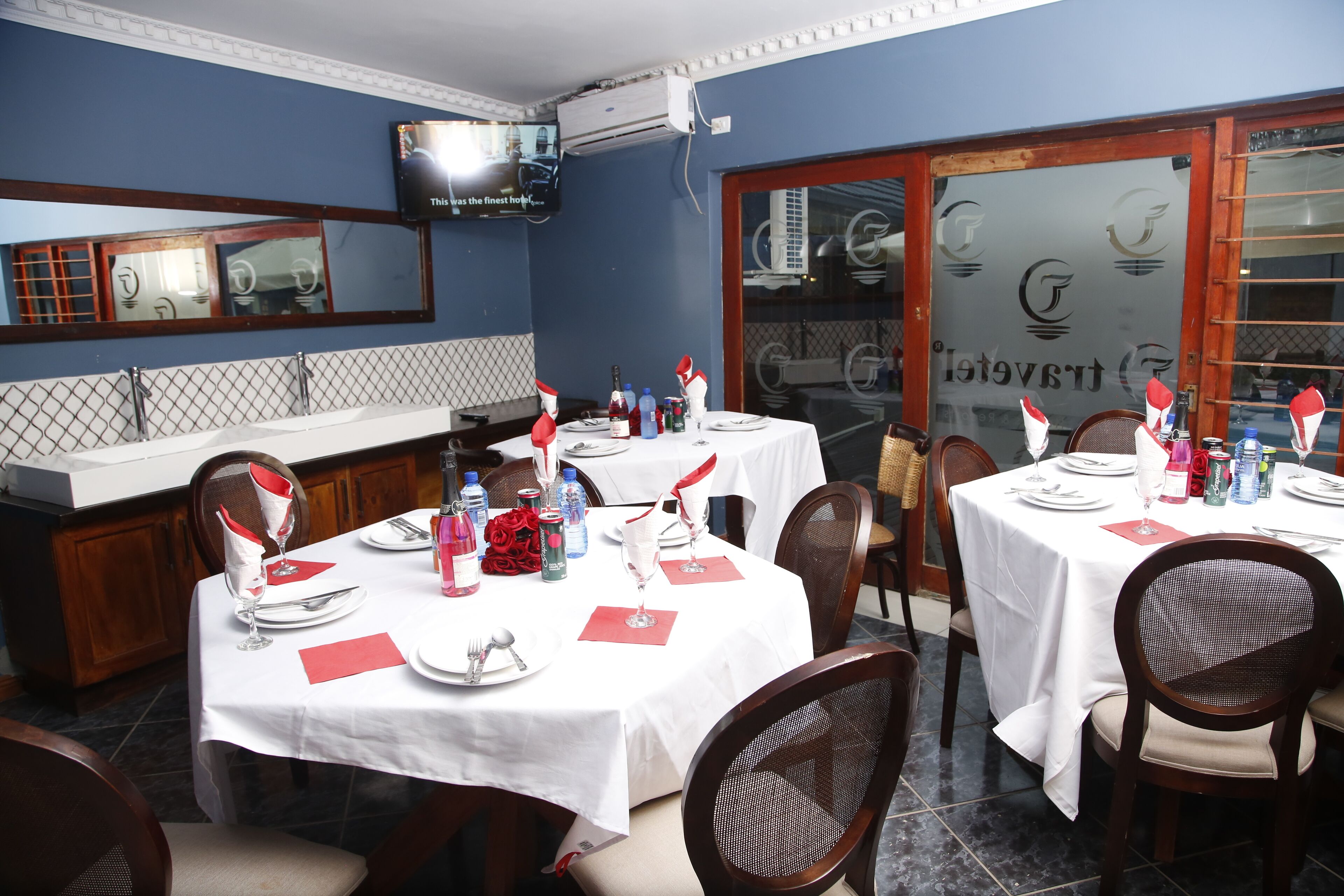 A dining area with tables set for two, each with white tablecloths, red napkins, and silverware. There are chairs with cushioned seats and backs. A television is mounted on the wall above the counter.