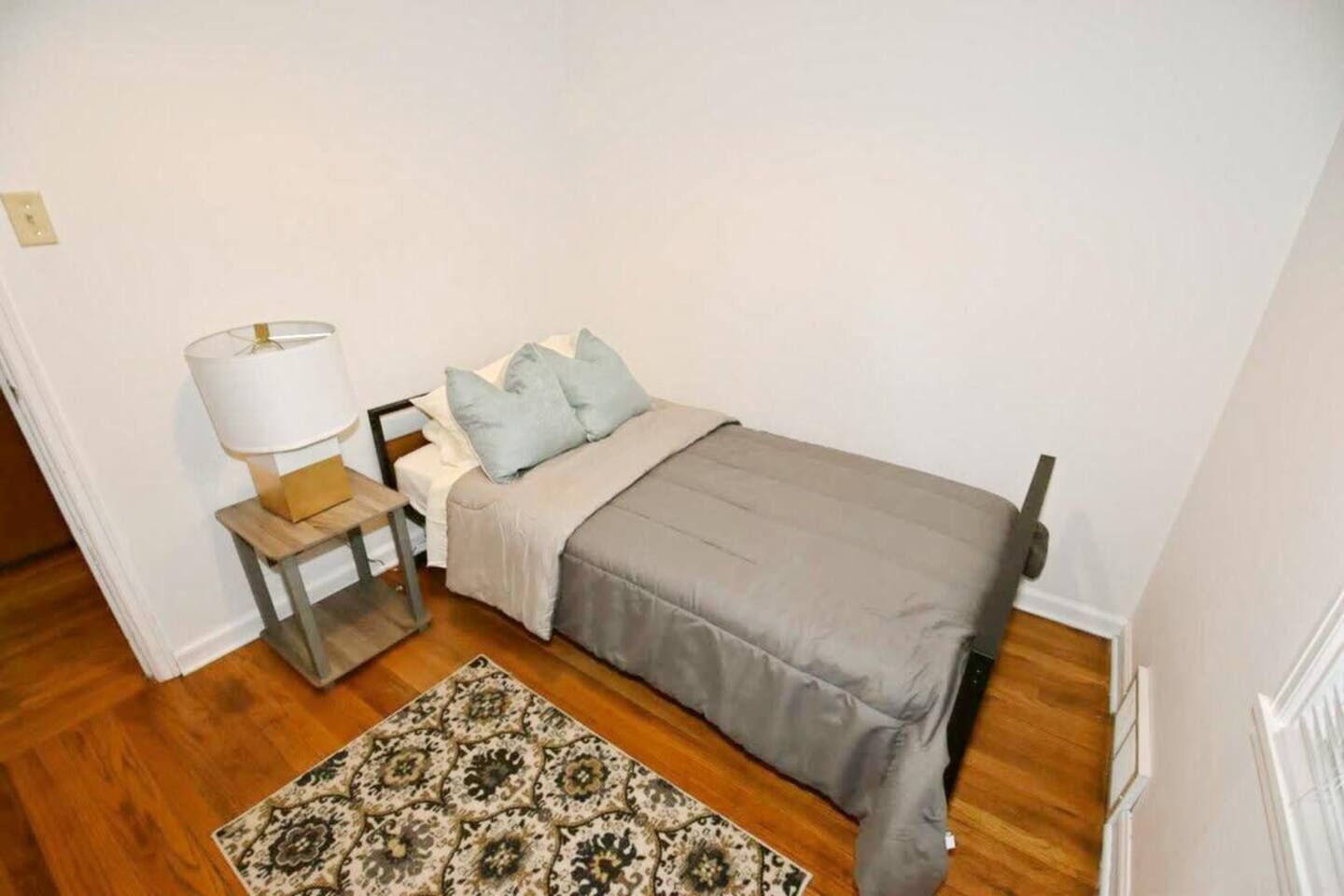 Una piccola camera con un letto singolo, pavimento in legno, un comodino con una lampada, un tappeto decorato e una coperta grigia.