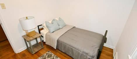 Una piccola camera con un letto singolo, pavimento in legno, un comodino con una lampada, un tappeto decorato e una coperta grigia.