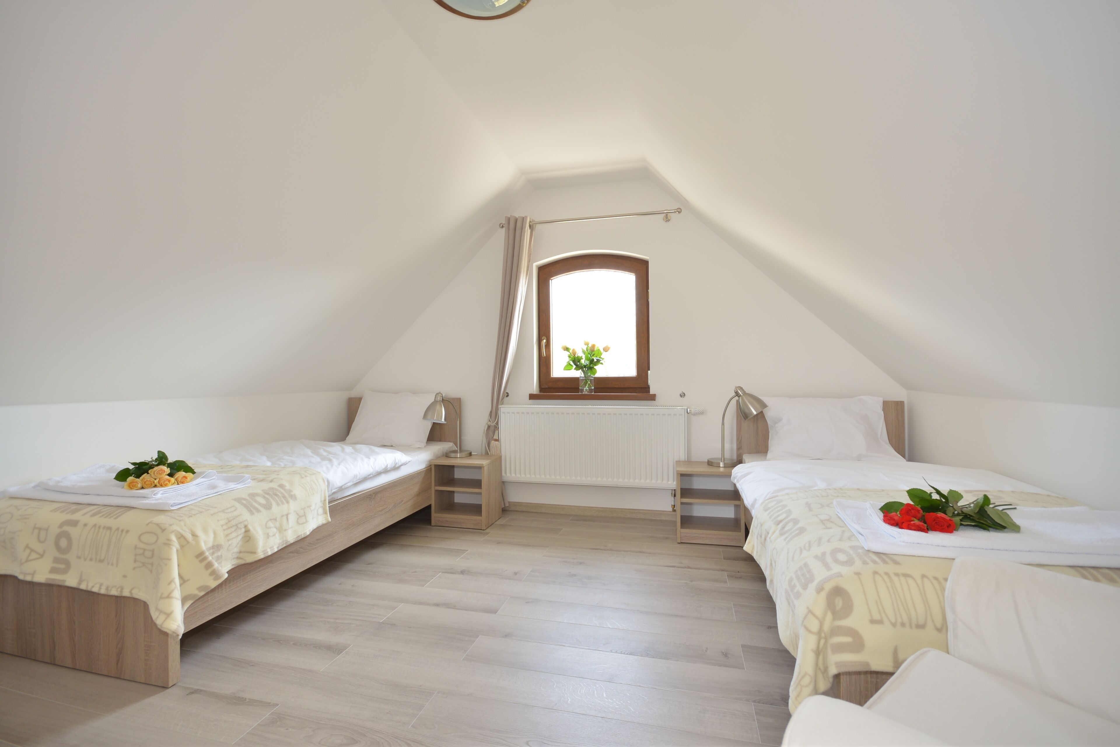 Une petite pièce bien éclairée, avec deux lits simples, une fenêtre avec des rideaux, un radiateur et une petite table avec une lampe.