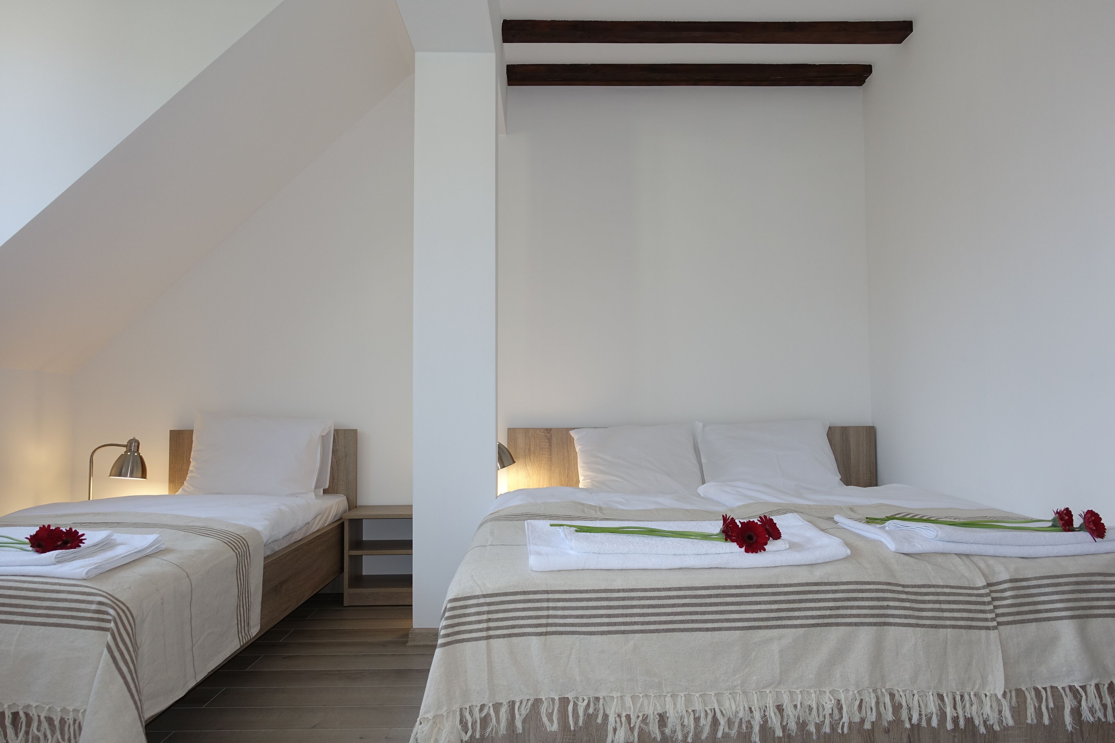A hotel room with two beds, each with a white bedspread and a wooden headboard. There is a nightstand with a lamp on the left bed, and towels with red flowers on the beds.