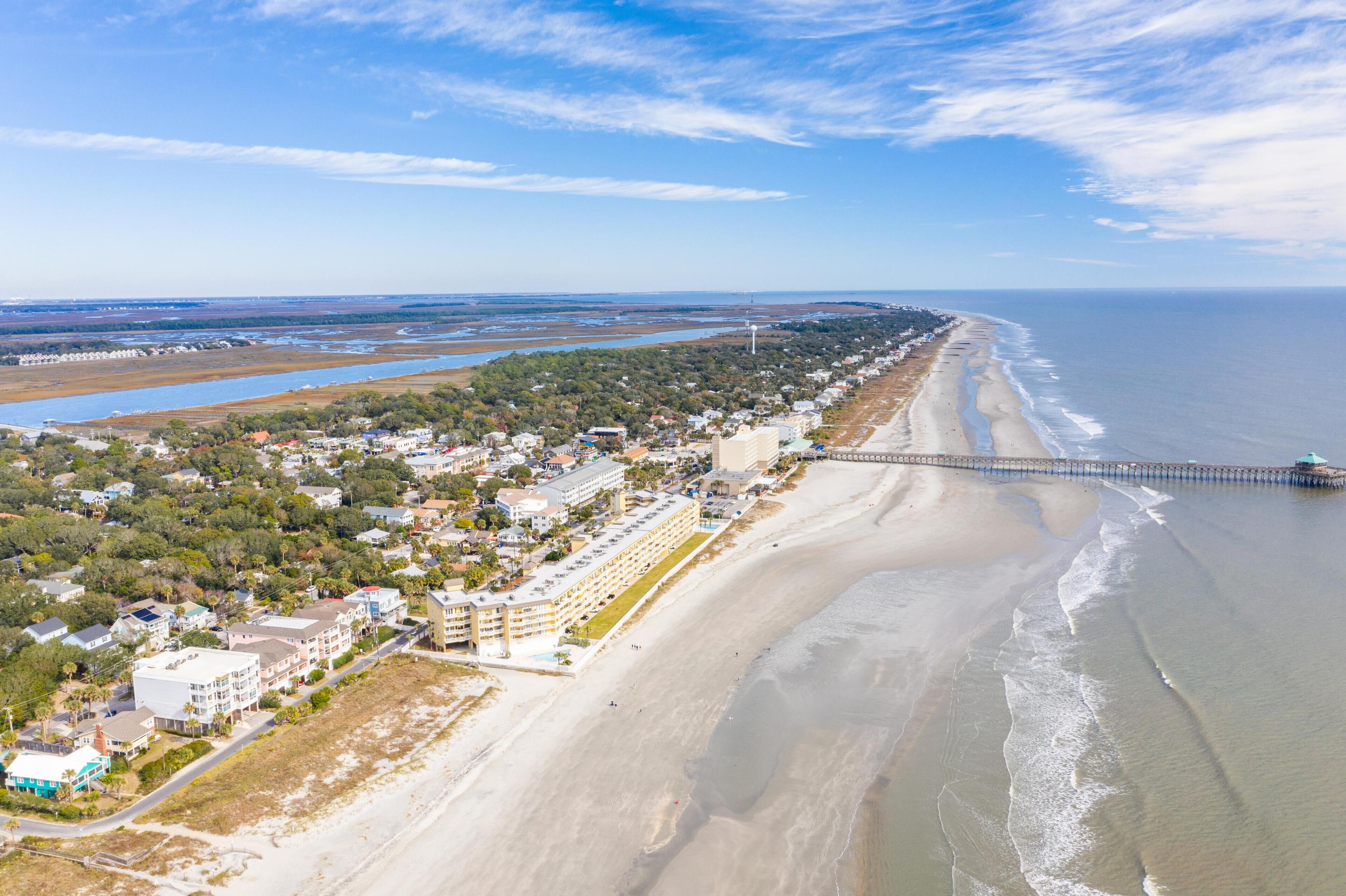 An aerial view of a coastal town with a long beachfront, a pier extending into the water, and a mix of residential and commercial buildings.