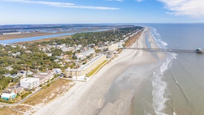 An aerial view of a coastal town with a long beachfront, a pier extending into the water, and a mix of residential and commercial buildings. - Oceanfront Condo w/ Dual Balconies + Walk to Folly Pier (Folly Beach)
