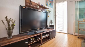 Una sala moderna con un televisor de pantalla plana grande sobre un mueble de madera, un florero con plantas secas y un cuadro colorido en la pared.