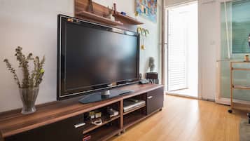 Una sala moderna con un televisor de pantalla plana grande sobre un mueble de madera, un florero con plantas secas y un cuadro colorido en la pared.