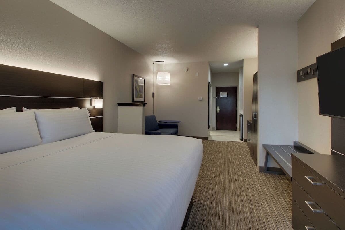 Ein Hotelzimmer mit einem großen Bett, einem Schreibtisch, einem Sessel, einem Fernseher und einem im Hintergrund sichtbaren Badezimmer.