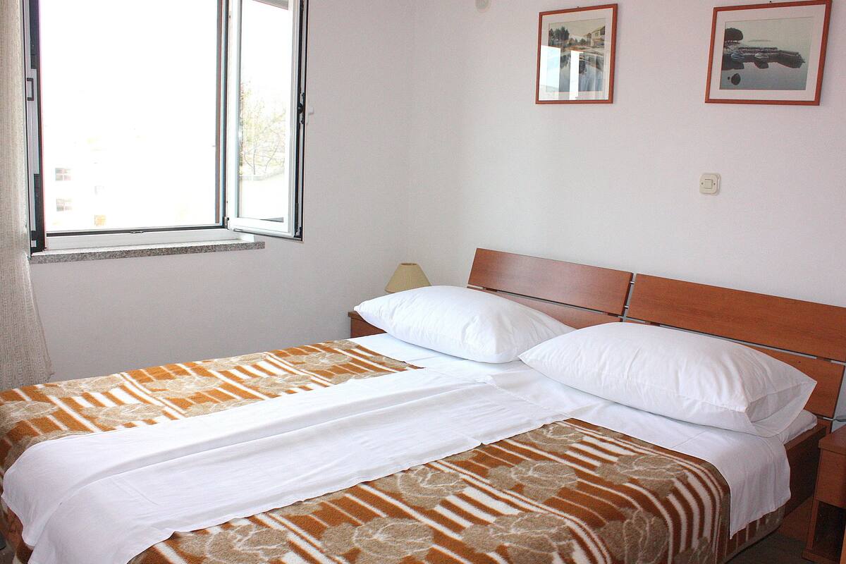 A neatly made bed with a patterned bedspread, a bedside table with a lamp, and a window with a view of buildings.