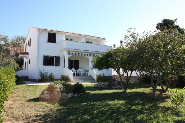 Um edifício branco de dois andares com telhado de telhas vermelhas, um alpendre coberto com cadeiras brancas e um jardim bem cuidado com diversas plantas e uma grande árvore.