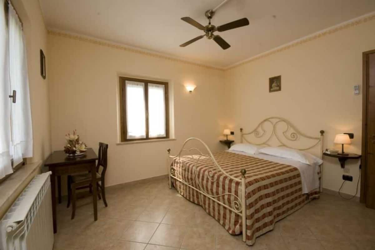 A bedroom with a single bed, a desk with a chair, a window, a ceiling fan, and two bedside lamps.