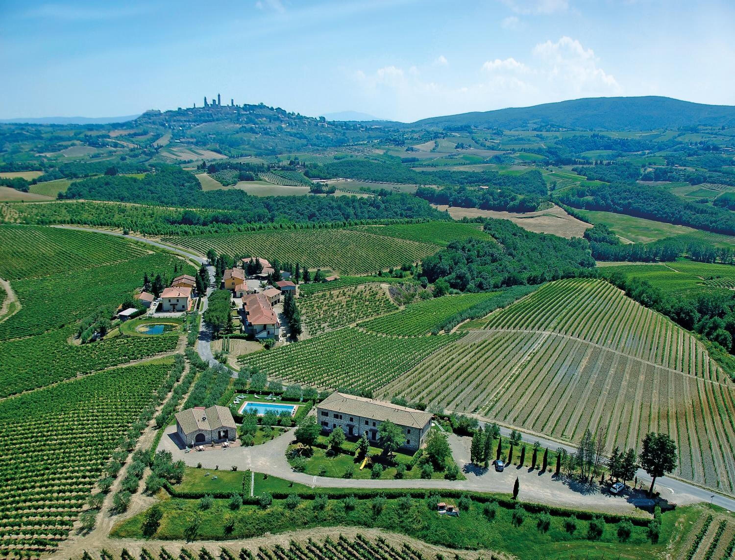 Une vue aérienne d’un vignoble avec des terrasses et un ensemble de bâtiments, dont une grande maison en pierre et des constructions plus petites, le tout entouré d’une végétation luxuriante et de collines ondulantes.