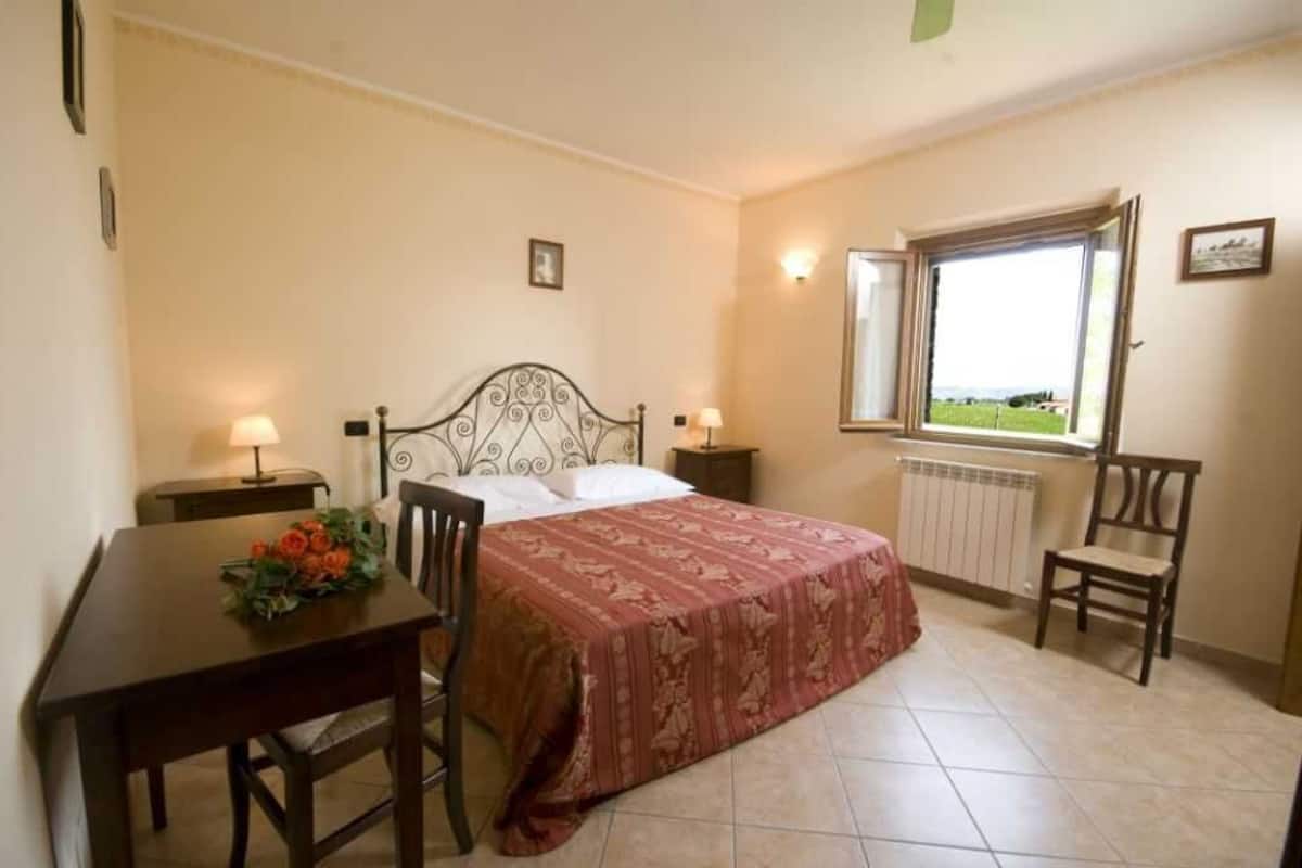 A bedroom with a bed, a table with a flower arrangement, a chair, a window with a view, and a radiator.
