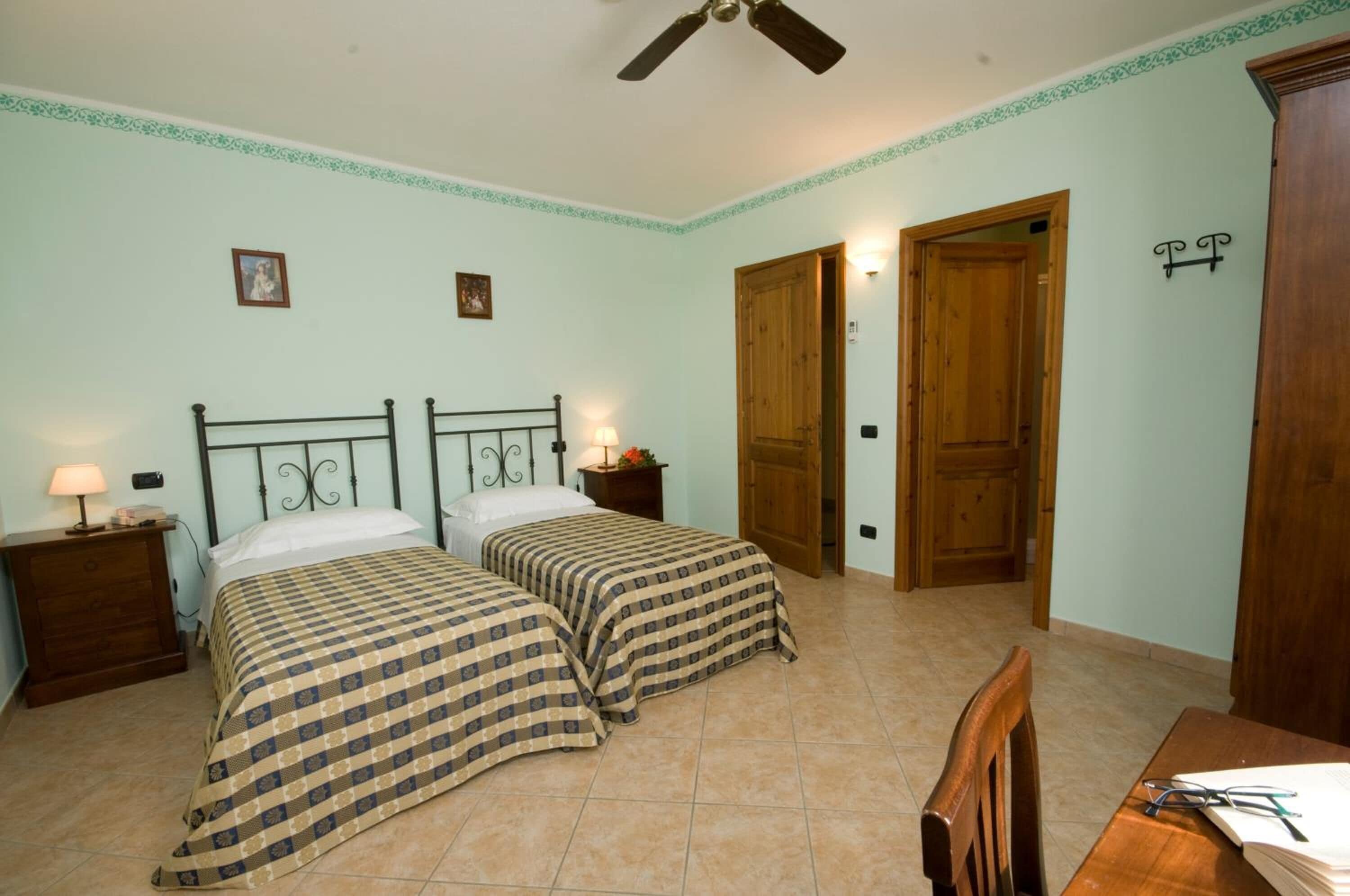 A bedroom with two beds, a wooden door, a desk with a chair, and a ceiling fan.