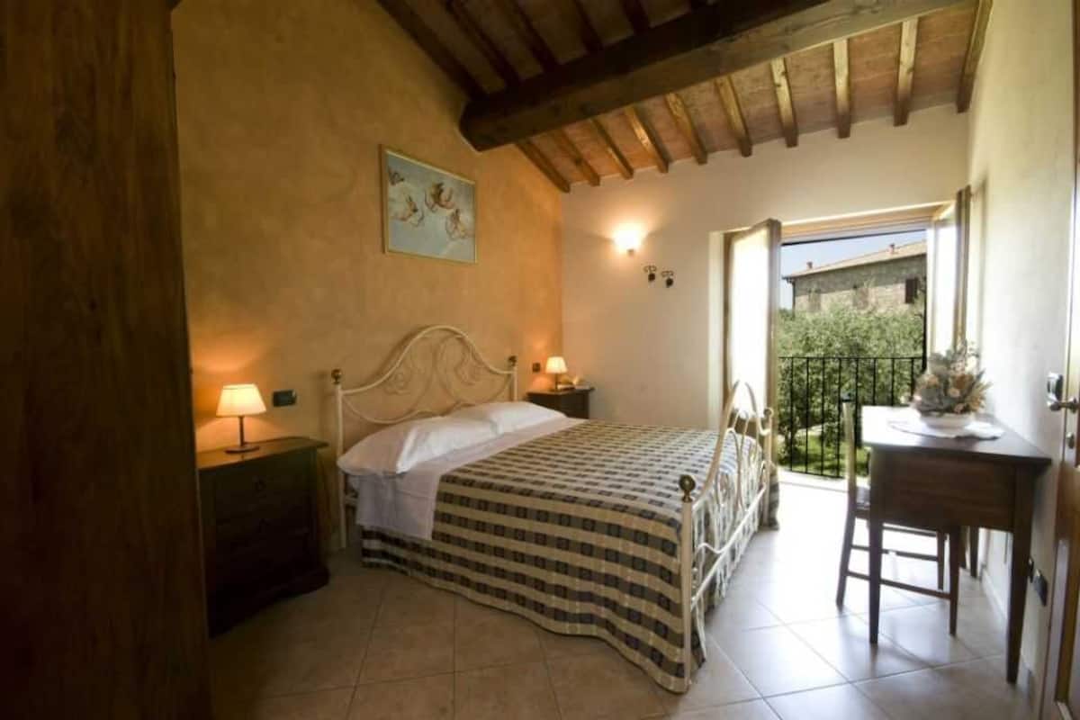 A bedroom with a bed, bedside lamps, a wooden ceiling, a painting on the wall, and a balcony with a table and chair.