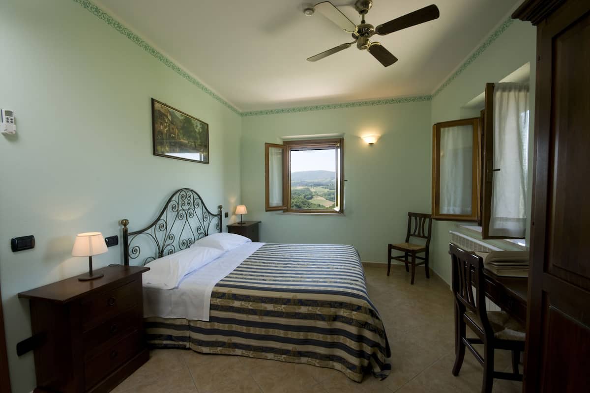 A bedroom with a bed, a desk, a chair, a window with a view, a ceiling fan, and a painting on the wall.