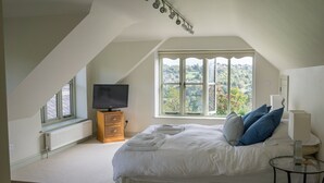 A bedroom with a large bed, a TV on a wooden stand, a window with a view of greenery, and a small table with a lamp. - The Retreat (Stroud)