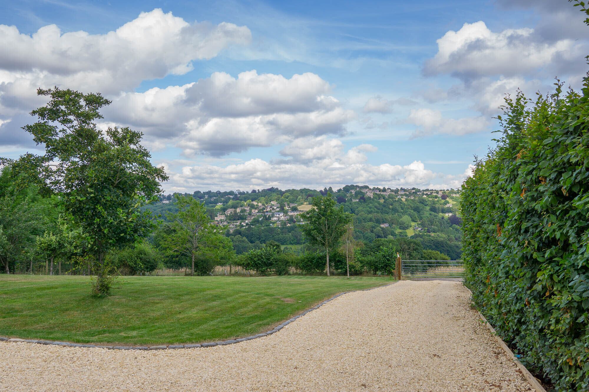 A gravel path leading through a grassy area with trees and a hedge on one side, and a hill with buildings in the distance.