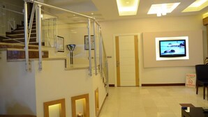 A modern hotel lobby with a glass staircase, a flat-screen TV mounted on the wall, and a sign indicating no smoking. - Napa Otel (Denizli)