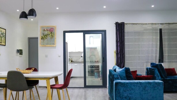A modern living room with a blue sofa, a dining table with chairs, and a kitchen visible through a glass door.