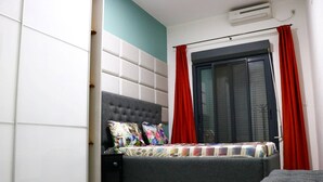 A small, compact bedroom with a single bed, a nightstand, a window with red curtains, and a wall-mounted air conditioner.